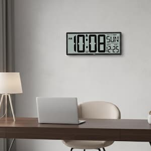 Könnte beinhalten: Eine rechteckige Digitaluhr mit schwarzem Rahmen zeigt die Uhrzeit 10:08 AM an, mit Tag, Datum und Temperatur. Die Uhr ist an einer weißen Wand über einem Schreibtisch mit Laptop und Lampe angebracht.