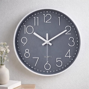 May include: A round wall clock with a gray face and white hands and numerals. The clock has a white frame and the word "QUARTZ" is printed at the bottom. The clock is mounted on a white wall.