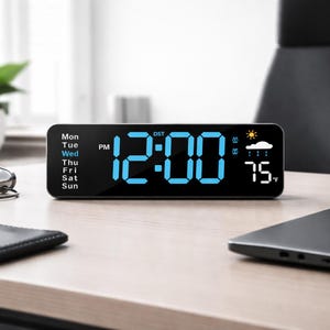 May include: A black digital alarm clock displaying the time as 12:00 PM in bright blue digits. The clock also shows the day of the week, weather icons, and a temperature of 75°F. The clock is rectangular with rounded corners.
