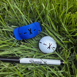 Pode incluir: Uma ferramenta de alinhamento de bola de golfe de plástico azul com uma letra X preta, uma bola de golfe branca marcada com uma letra X preta e linhas horizontais, e um marcador permanente Sharpie Ultra Fine Point preto e branco.