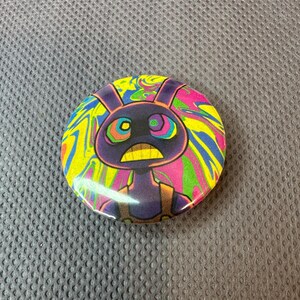 May include: A circular badge featuring a cartoon rabbit character. The rabbit is depicted in purple with a yellow mouth and a startled expression. The background has a swirling pattern of yellow, pink, green, and blue.