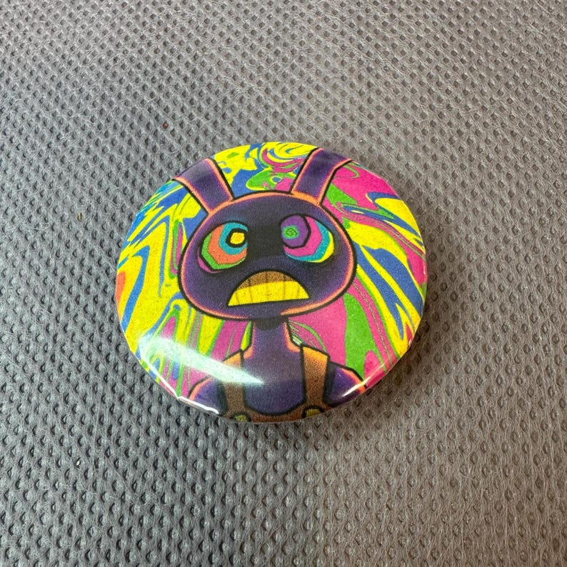 May include: A circular badge featuring a cartoon rabbit character. The rabbit is depicted in purple with a yellow mouth and a startled expression. The background has a swirling pattern of yellow, pink, green, and blue.