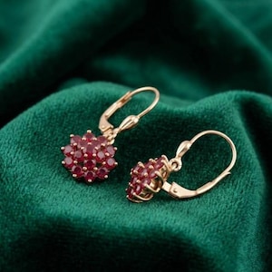 May include: A pair of gold-colored earrings with a flower-like design. Each earring features a cluster of small, round, red gemstones. The earrings are set against a dark green, textured background.