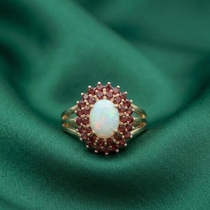 May include: A gold ring featuring a large oval opal center stone surrounded by a halo of small, round, red gemstones. The ring has a double band design. The background is a dark green fabric.