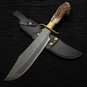 May include: A large hunting knife with a textured blade and a handle made of antler. The knife has a brass bolster and is paired with a black leather sheath. The knife is designed for outdoor use.