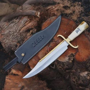 Puede incluir: Un cuchillo grande con una hoja plateada, una guarda de latón y un mango color crema. El cuchillo se muestra con una funda de cuero negro, cosida con hilo blanco. El cuchillo y la funda descansan sobre una superficie de madera marrón texturizada.