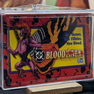 May include: A DC Bloodlines trading card in a clear plastic case. The card features a pink and purple alien-like figure with a butterfly wing design. The card has the text "BLOODLINES" and "New Heroes. New Villains. New Blood."