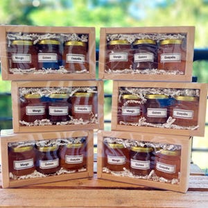 May include: Six wooden gift boxes, each containing three small glass jars of preserves. The jars have gold lids and white labels that read "Mango", "Guineo", and "Guayaba". The jars are nestled in shredded paper.