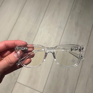 May include: Clear rectangular eyeglasses with silver detailing on the temples and hinges. The lenses are clear, and the frame is transparent. A silver emblem is visible on the right lens.