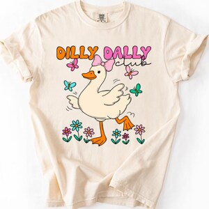 May include: A cream-colored short-sleeve t-shirt featuring a graphic of a white duck wearing a pink bow. The text DILLY DALLY CLUB is printed above the duck. The design includes colorful butterflies and flowers in shades of pink, orange, and teal.