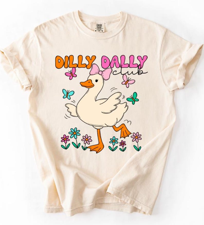 May include: A cream-colored short-sleeve t-shirt featuring a graphic of a white duck wearing a pink bow. The text DILLY DALLY CLUB is printed above the duck. The design includes colorful butterflies and flowers in shades of pink, orange, and teal.