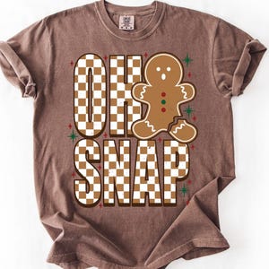 May include: A brown short-sleeve t-shirt featuring a graphic with the text "OH SNAP" in a white and brown checkered pattern. Next to the text is a brown gingerbread person with a broken leg, accented by small red and green star shapes.