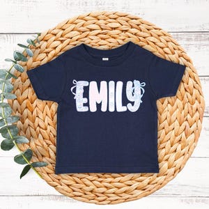 May include: Navy blue toddler t-shirt with the name "EMILY" in white, light blue, and pink lettering. The name is decorated with floral and bow accents. The shirt is displayed on a woven, circular placemat.