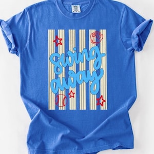 May include: A blue t-shirt with a baseball-themed design. The shirt features the words "Swing Away" in a light blue script, along with baseball-related graphics such as a baseball, glove, and stars. Vertical cream stripes are behind the text.