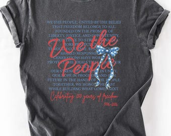 May include: Dark gray t-shirt with a patriotic design. The shirt features the words "We the People" in red script, along with a blue bow with white stars. The text celebrates 250 years of freedom, from 1776 to 2026.