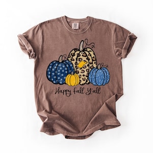 May include: A brown short-sleeve t-shirt featuring a graphic of four pumpkins in blue polka dot, leopard print, solid yellow, and blue patterns. The leopard print pumpkin includes a small yellow state outline. The text reads Happy fall Y'all.