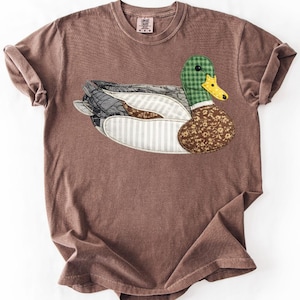 May include: A brown short-sleeved t-shirt featuring a large, central appliqué of a mallard duck. The duck is constructed from various fabric patches, including green gingham, yellow, brown floral, and striped patterns, with a black button eye.