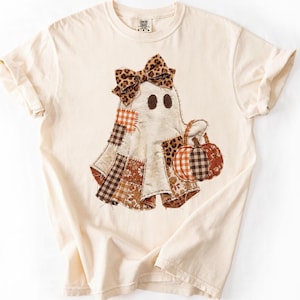 May include: A cream-colored short-sleeve t-shirt featuring a central graphic of a ghost wearing a leopard print bow. The ghost is decorated with various fabric patches in plaid, floral, and leopard patterns and holds a pumpkin bucket.
