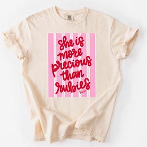 May include: Cream-colored t-shirt with rolled sleeves. The shirt features a pink and white striped design with the text "She is more precious than rubies" in red script. The shirt is a casual, comfortable style.
