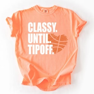 May include: A coral-colored t-shirt with the text "CLASSY. UNTIL. TIPOFF." in white block letters. A basketball design is incorporated into the word "TIPOFF" in the shape of a heart. The shirt has short sleeves and a crew neck.