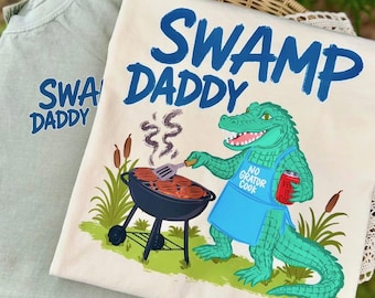 May include: A cream-colored t-shirt featuring a graphic of a green alligator wearing a blue apron that says NO GRATOR COOK, holding a red drink, and grilling burgers. The text SWAMP DADDY is printed in blue above the alligator. A sage green shirt is underneath.