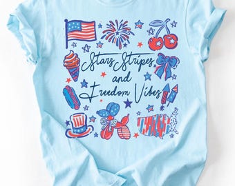 May include: Light blue t-shirt with a patriotic design. The shirt features illustrations of an American flag, fireworks, cherries, an ice cream cone, a hot dog, a top hat, a balloon dog, a bow, a map of the USA, and the text "Stars Stripes and Freedom Vibes."