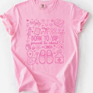 May include: A light pink t-shirt with a pink graphic design featuring medical-themed illustrations and the text "BORN TO YAP forced to chart". The design includes a stethoscope, shoes, and a thermometer.