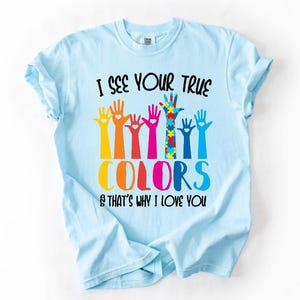 May include: Light blue t-shirt with the text "I SEE YOUR TRUE COLORS & THAT'S WHY I LOVE YOU." The shirt features colorful hand graphics, including a hand with a puzzle piece design.