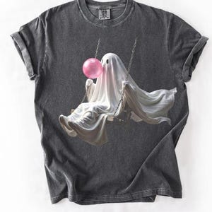 May include: A charcoal gray short-sleeve t-shirt featuring a graphic print of a white ghost draped in a sheet, sitting on a swing and blowing a large pink bubblegum bubble. The ghost has two simple black eye holes and is suspended by metal chains.