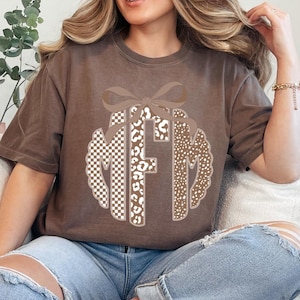 May include: Brown t-shirt featuring a monogram design with the letters "MFM". The monogram is decorated with a bow and a mix of patterns, including checkered, leopard print, and dotted designs. The shirt is a neutral color and has a relaxed fit.