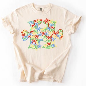 May include: Cream-colored t-shirt featuring a colorful puzzle piece design. The puzzle piece is filled with a pattern of bows in red, blue, green, and yellow. The shirt has short sleeves and a crew neck.