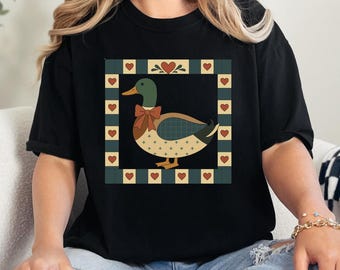 May include: Black t-shirt featuring a duck design. The duck has a green head, a brown body with a cross pattern, and a red bow. The design is framed by a square with hearts and a green background. The shirt is a casual style.