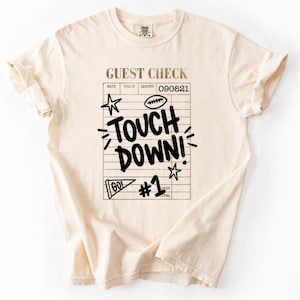 May include: A cream-colored short-sleeve t-shirt featuring a black graphic print of a restaurant guest check. The design includes the text GUEST CHECK, TOUCH DOWN!, GO!, and #1, along with illustrations of a football and two stars.