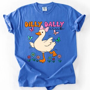 May include: A blue short-sleeve t-shirt featuring a graphic of a white duck wearing a pink bow. The text DILLY DALLY club is printed above the duck. The design includes colorful butterflies and flowers in shades of pink, orange, blue, and green.