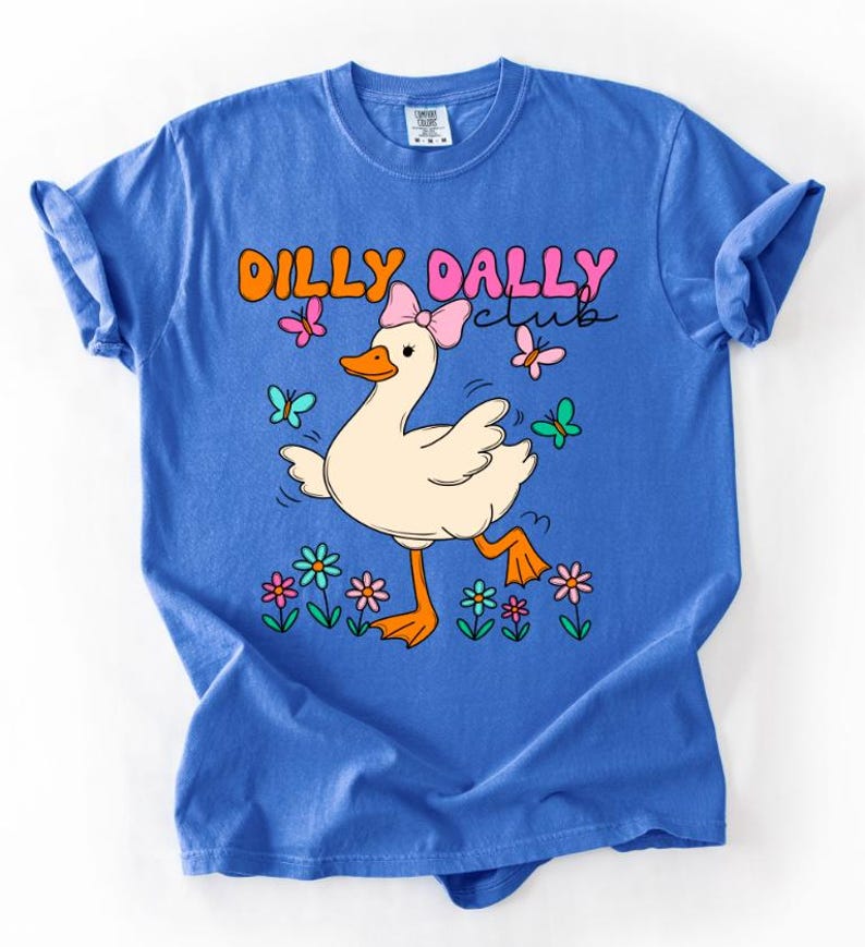May include: A blue short-sleeve t-shirt featuring a graphic of a white duck wearing a pink bow. The text DILLY DALLY club is printed above the duck. The design includes colorful butterflies and flowers in shades of pink, orange, blue, and green.