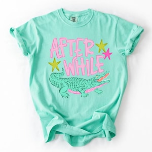 May include: A turquoise t-shirt with rolled-up sleeves featuring the text "AFTER WHILE" in pink, along with a green alligator illustration and star accents. The shirt is made of a soft material.