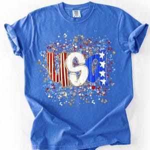 May include: A bright blue short-sleeve t-shirt featuring a central graphic with the letters USA. The U has red and white stripes, the S is white with a distressed texture, and the A is blue with white stars, surrounded by a colorful paint splatter.