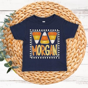 May include: A navy blue short-sleeve t-shirt featuring a graphic print of three candy corn pieces above the name MORGAN in orange and yellow letters, all enclosed within a black and white checkered square border on the front of the shirt.