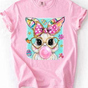 May include: Pink t-shirt featuring a cartoon bunny with a pink bubblegum bubble. The bunny wears heart-shaped glasses and a polka-dot bow. Easter eggs and flowers surround the bunny on a light blue background.