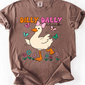 May include: A brown short-sleeve t-shirt featuring a graphic of a white duck wearing a pink bow. The text DILLY DALLY club is printed above the duck, surrounded by colorful butterflies and flowers in shades of pink, orange, blue, and green.