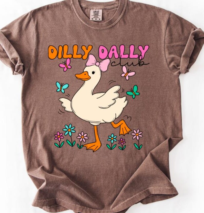 May include: A brown short-sleeve t-shirt featuring a graphic of a white duck wearing a pink bow. The text DILLY DALLY club is printed above the duck, surrounded by colorful butterflies and flowers in shades of pink, orange, blue, and green.