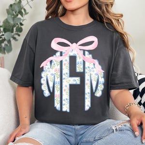 May include: Dark gray t-shirt featuring a floral monogram design with the letters MFM. The monogram is surrounded by a scalloped edge and topped with a pink bow. The floral pattern includes blue and pink flowers.