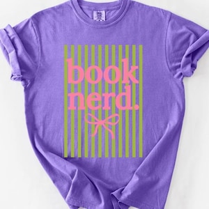 May include: A purple t-shirt with the words "book nerd." in pink, set against a green striped background. A pink bow is below the text. The shirt has a crew neck and short sleeves.