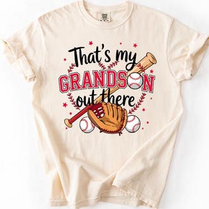 May include: Cream-colored t-shirt with the text "That's my GRANDSON out there" in black and red. The design includes a baseball bat, glove, and two baseballs. The shirt is a casual wear item.