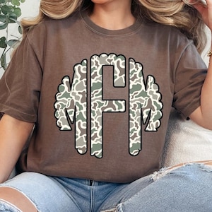 May include: A brown t-shirt featuring a monogram design with the letters "MFM" in a camouflage pattern. The letters are outlined in black and set against a brown background. The shirt has short sleeves and a relaxed fit.
