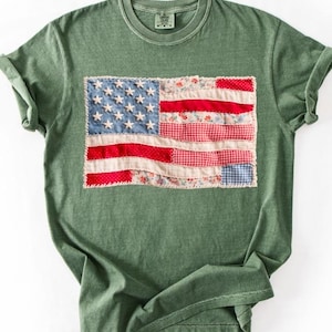 May include: A green t-shirt featuring a patchwork American flag design. The flag includes blue stars, red stripes, and floral and gingham patterns. The shirt has a crew neck and short sleeves.