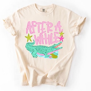 May include: Cream-colored t-shirt featuring a graphic of a green alligator with pink accents and the text "AFTER A WHILE" in pink. The design includes green and pink stars. The shirt has short sleeves and a crew neck.