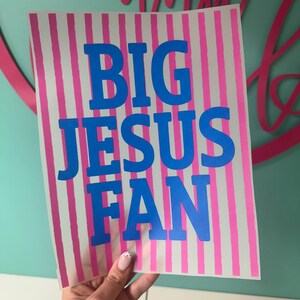 K&ouml;nnte beinhalten: Ein rechteckiges Schild mit vertikalen rosa und wei&szlig;en Streifen. Fettgedruckter blauer Text lautet "BIG JESUS FAN". Das Schild wird von einer Hand mit einem hellrosa und wei&szlig;en Nageldesign gehalten. Der Hintergrund ist hellt&uuml;rkis.