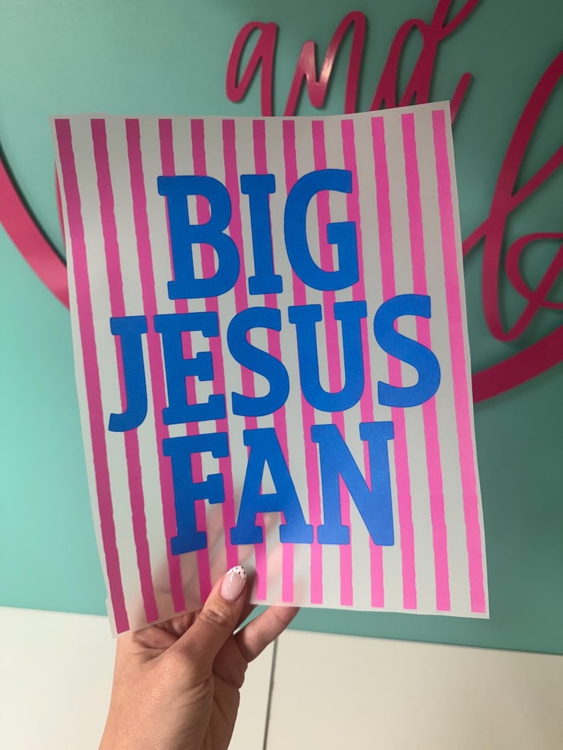 K&ouml;nnte beinhalten: Ein rechteckiges Schild mit vertikalen rosa und wei&szlig;en Streifen. Fettgedruckter blauer Text lautet "BIG JESUS FAN". Das Schild wird von einer Hand mit einem hellrosa und wei&szlig;en Nageldesign gehalten. Der Hintergrund ist hellt&uuml;rkis.