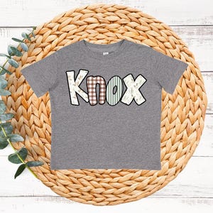 May include: A gray t-shirt with the name "Knox" in a decorative font. The letters are outlined in black and filled with various patterns, including plaid, stripes, and floral designs. The shirt is displayed on a woven, circular mat.